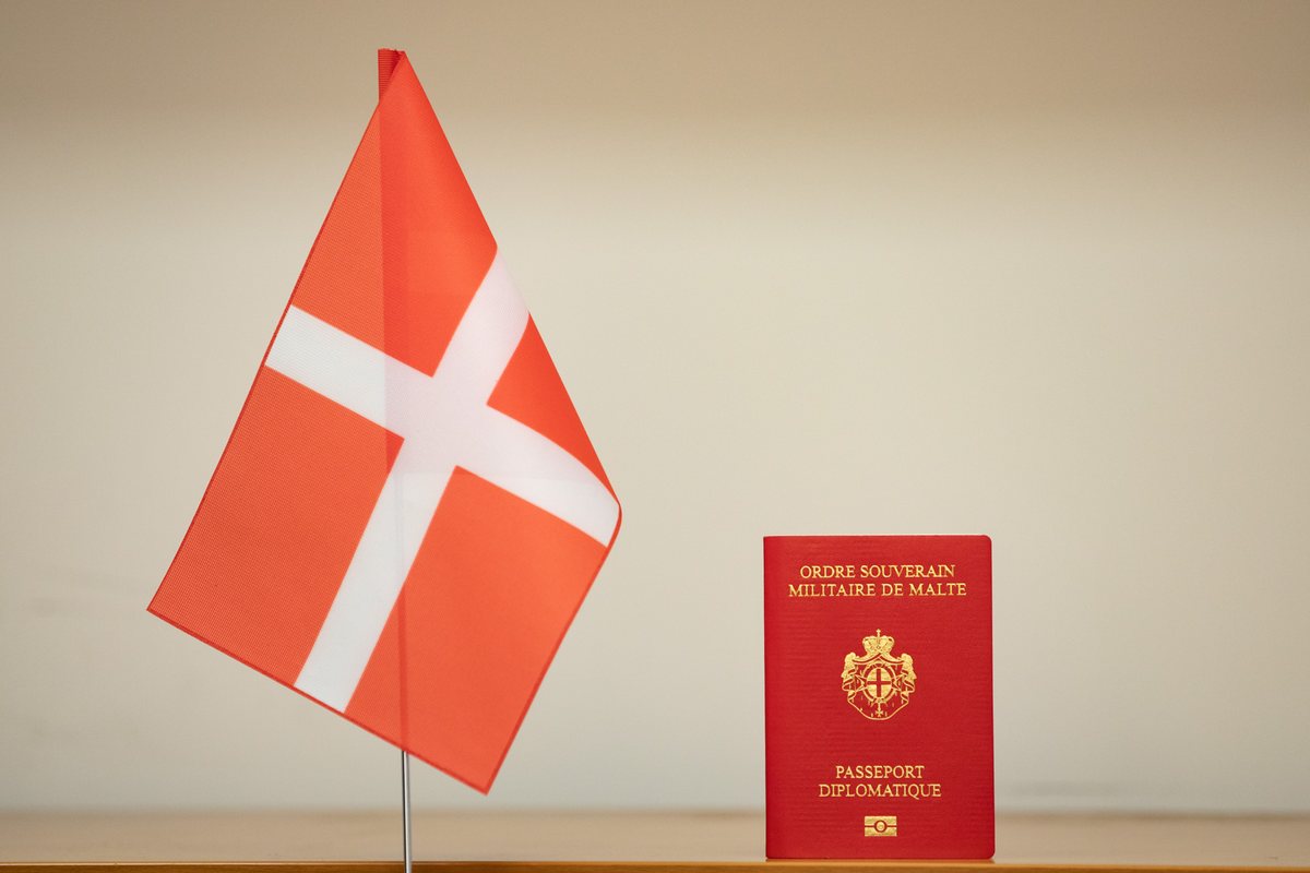 Red diplomatic passport of the Sovereign Military Order of Malta beside the Order’s flag.